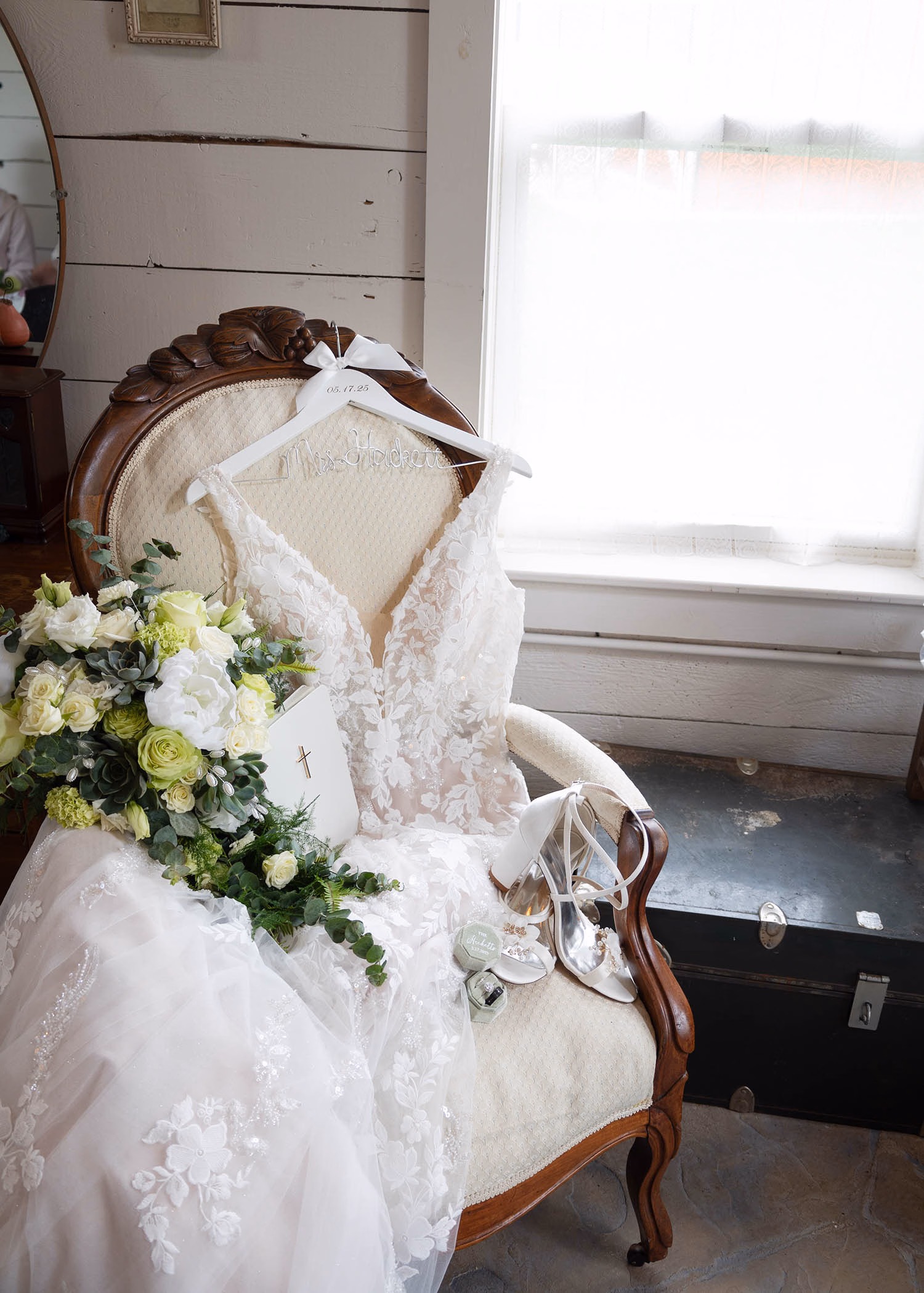 A vintage armchair holding a lace wedding gown, silver bridal shoes, and a white bible, captured by La Crosse, Wisconsin wedding photographer Captured by Nancy Christine Photography.