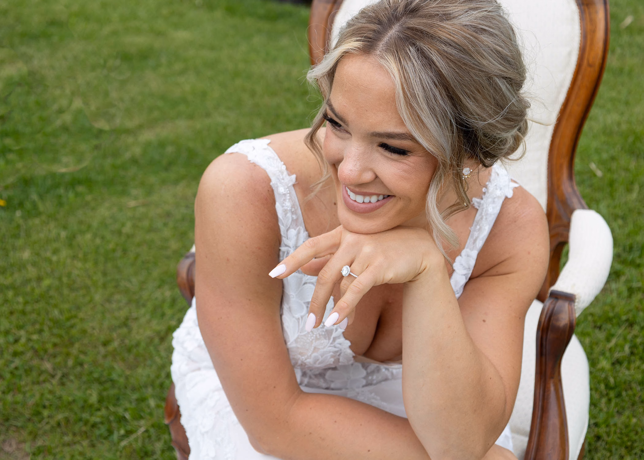 A smiling bride in her lace wedding dress leans forward to showcase her diamond engagement ring, captured by La Crosse, Wisconsin wedding photographer Captured by Nancy Christine Photography.