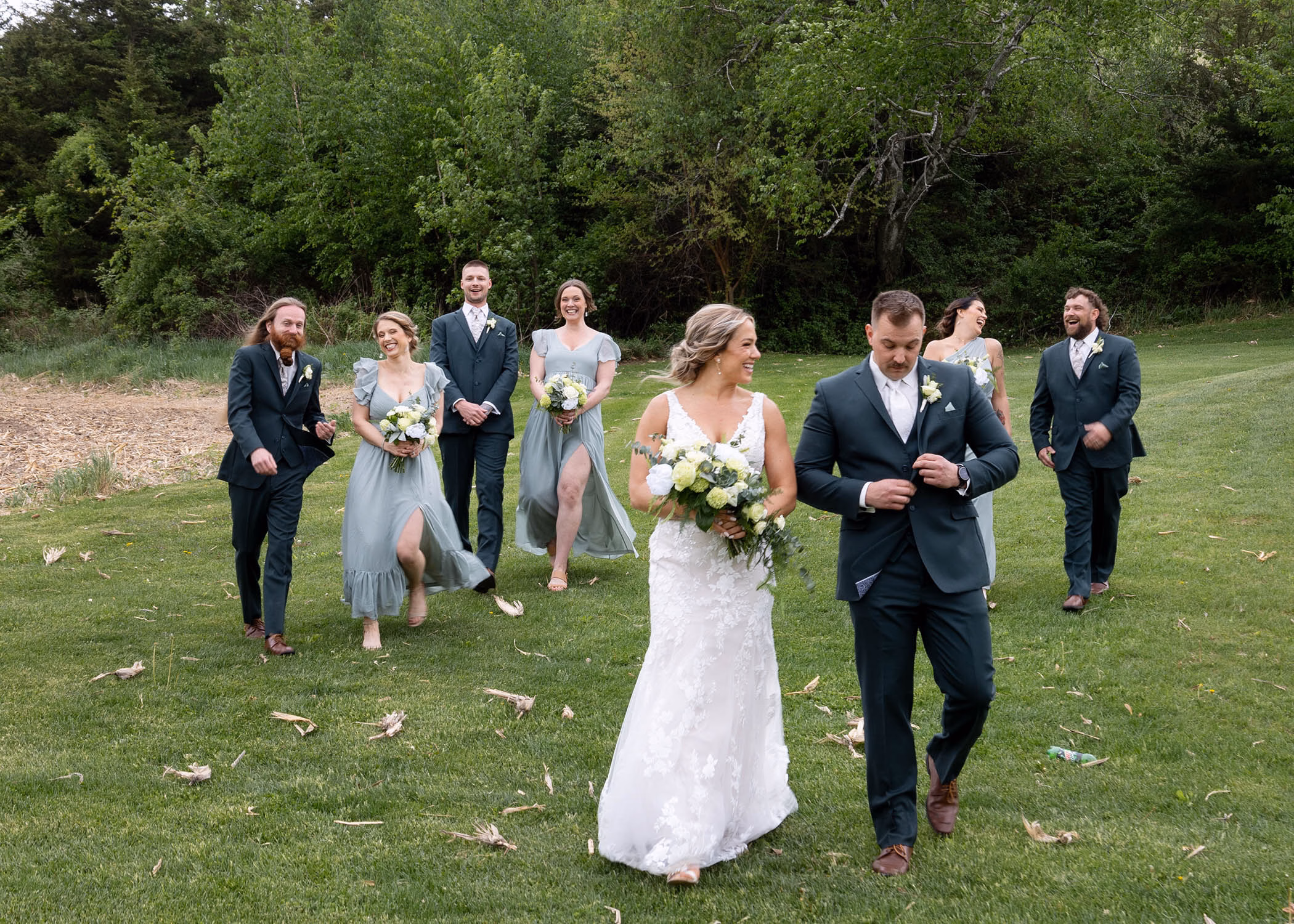 A joyful bride and groom walk across a grassy lawn with their bridal party laughing behind them, captured by La Crosse, Wisconsin wedding photographer Captured by Nancy Christine Photography.