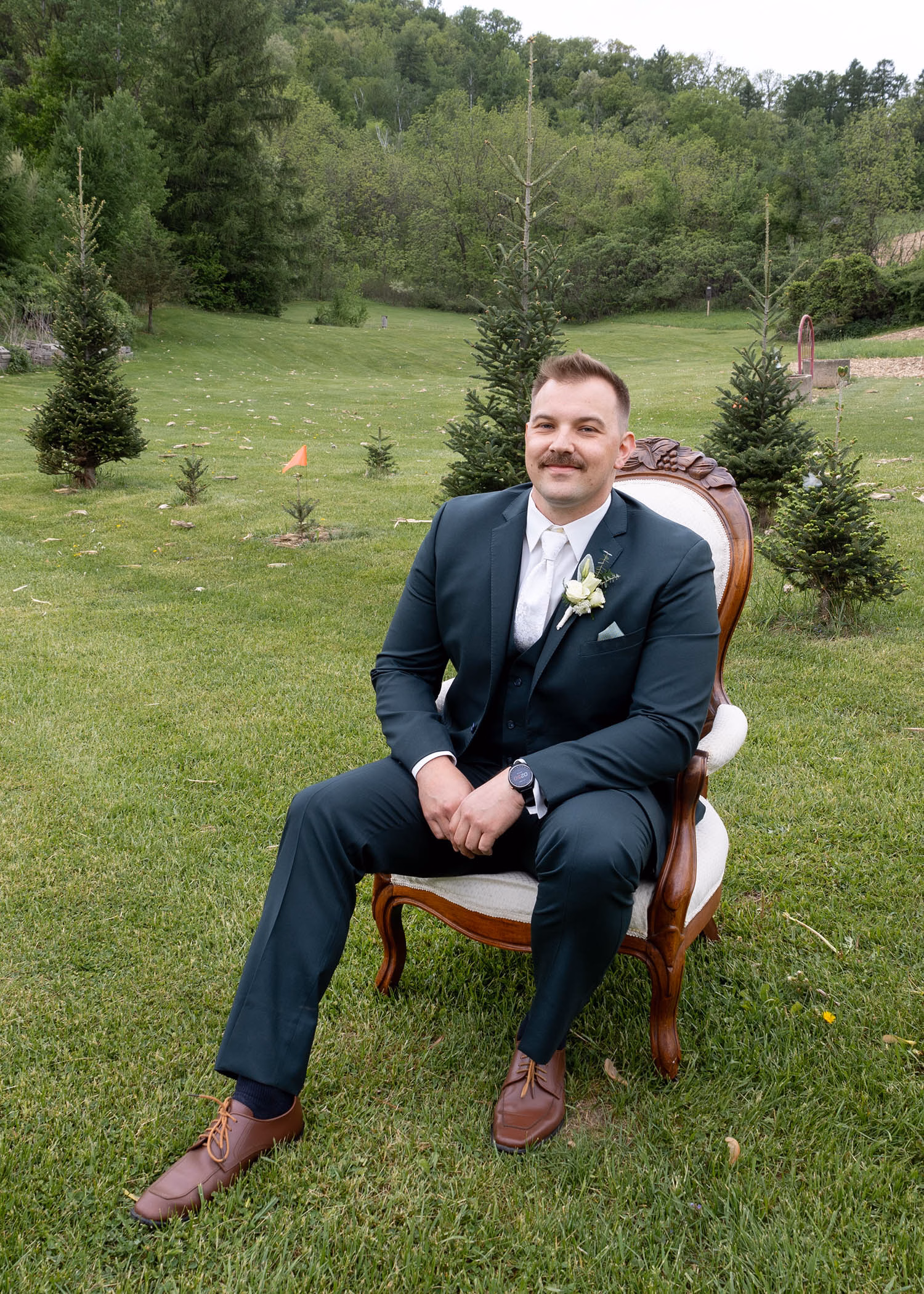 A portrait of a groom in a dark slate suit sitting on a vintage armchair in an outdoor grassy field, captured by La Crosse, Wisconsin wedding photographer Captured by Nancy Christine Photography.