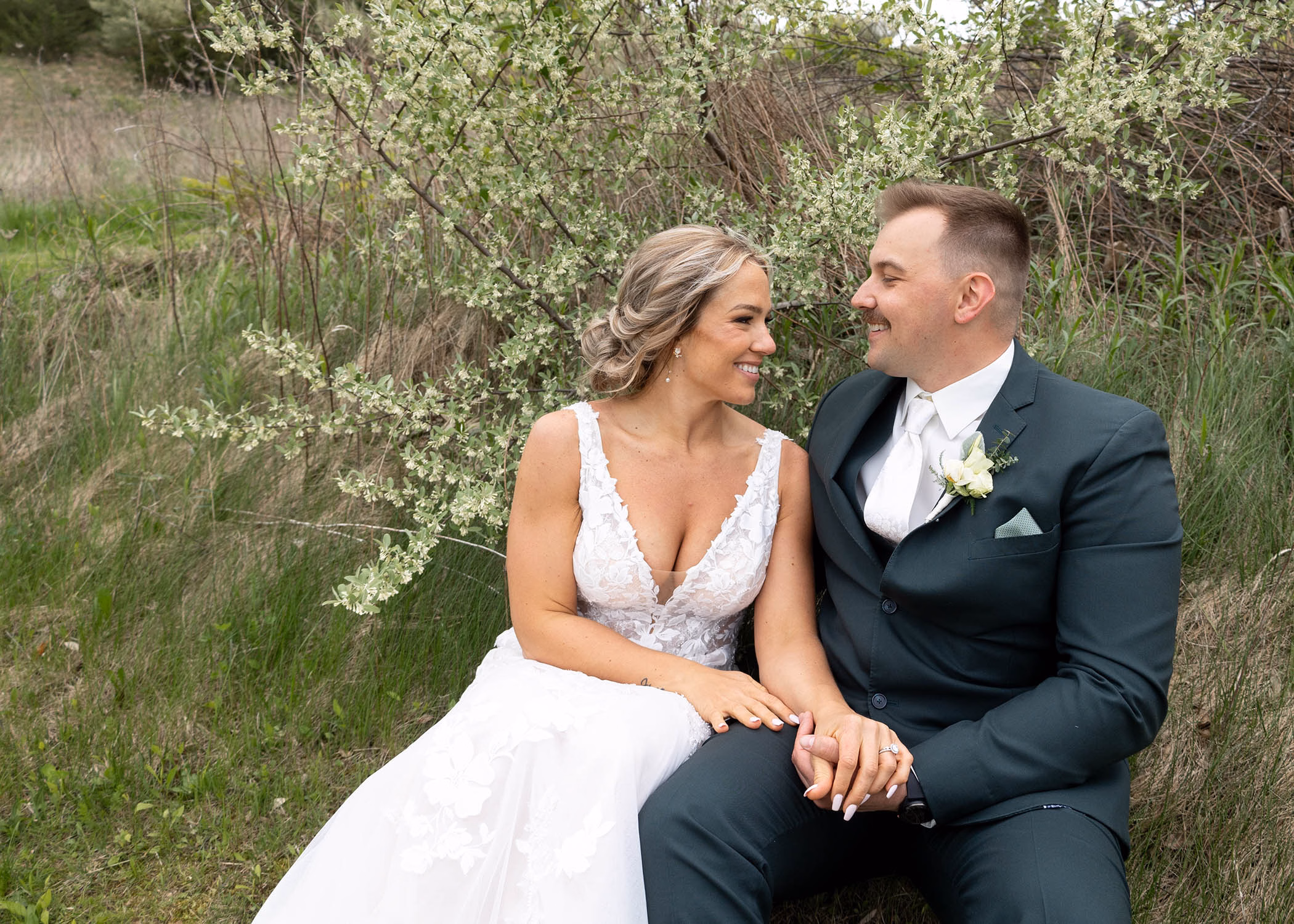 A bride and groom sitting together on a grassy hillside in front of a blooming white floral bush, captured by La Crosse, Wisconsin wedding photographer Captured by Nancy Christine Photography.