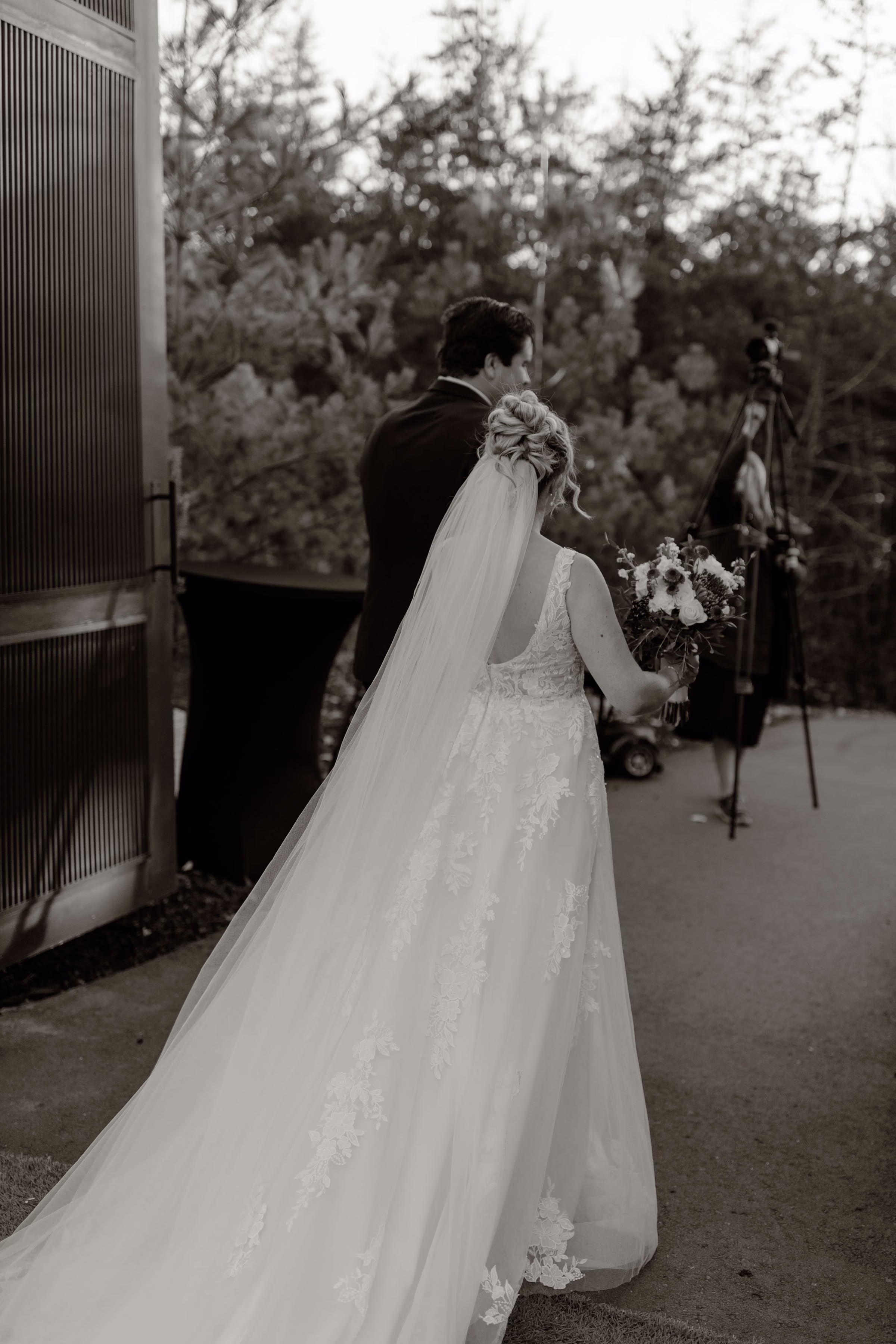 Black and white photograph captured from behind a bride and groom during a wedding coordinated by Rooted Weddings & Events. The bride wears an elegant, lace-detailed wedding gown with a long, flowing tulle veil and holds a rustic floral bouquet, while the groom walks beside her in a dark suit toward an outdoor wooded area.