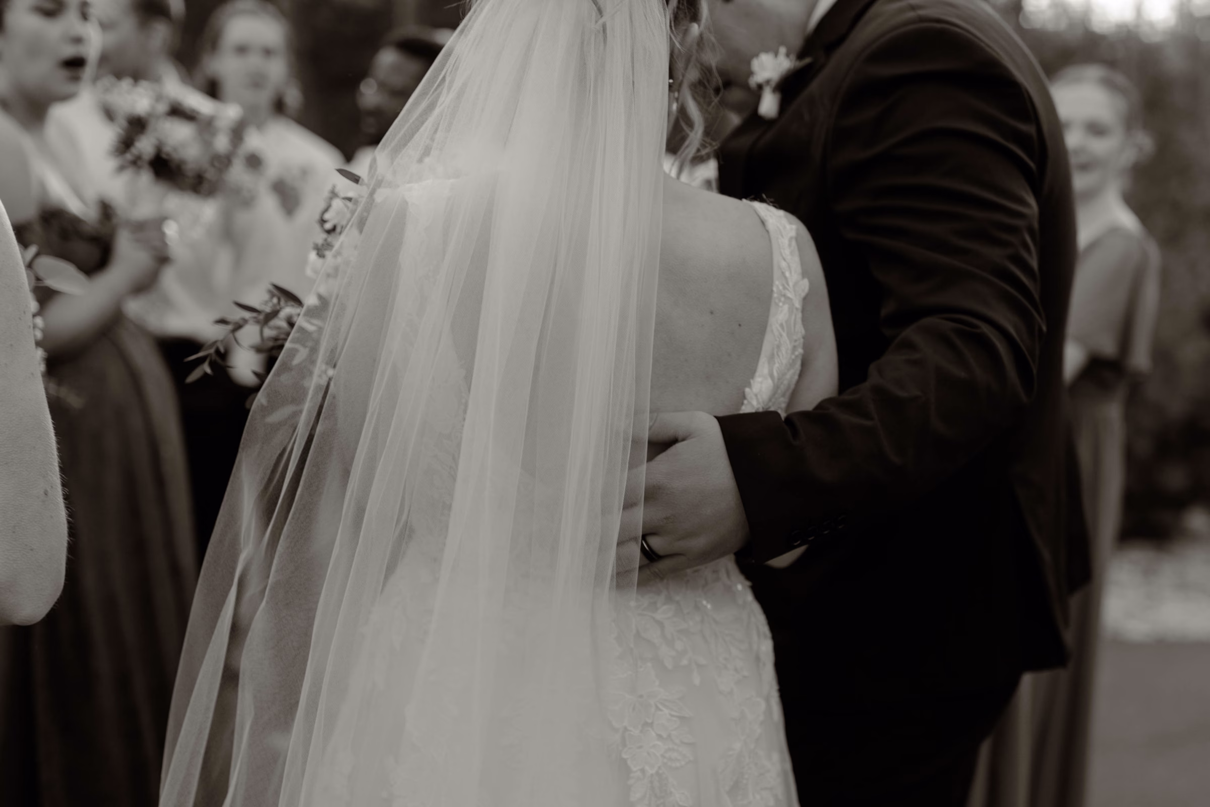 A black and white, close-up photograph captured from behind a bride and groom during a wedding coordinated by Rooted Weddings & Events. The groom wears a dark suit with a boutonniere and places his hand gently on the bride’s back, while the bride stands in an elegant, V-back lace wedding gown with a long tulle veil, surrounded by wedding party members softly blurred in the background.