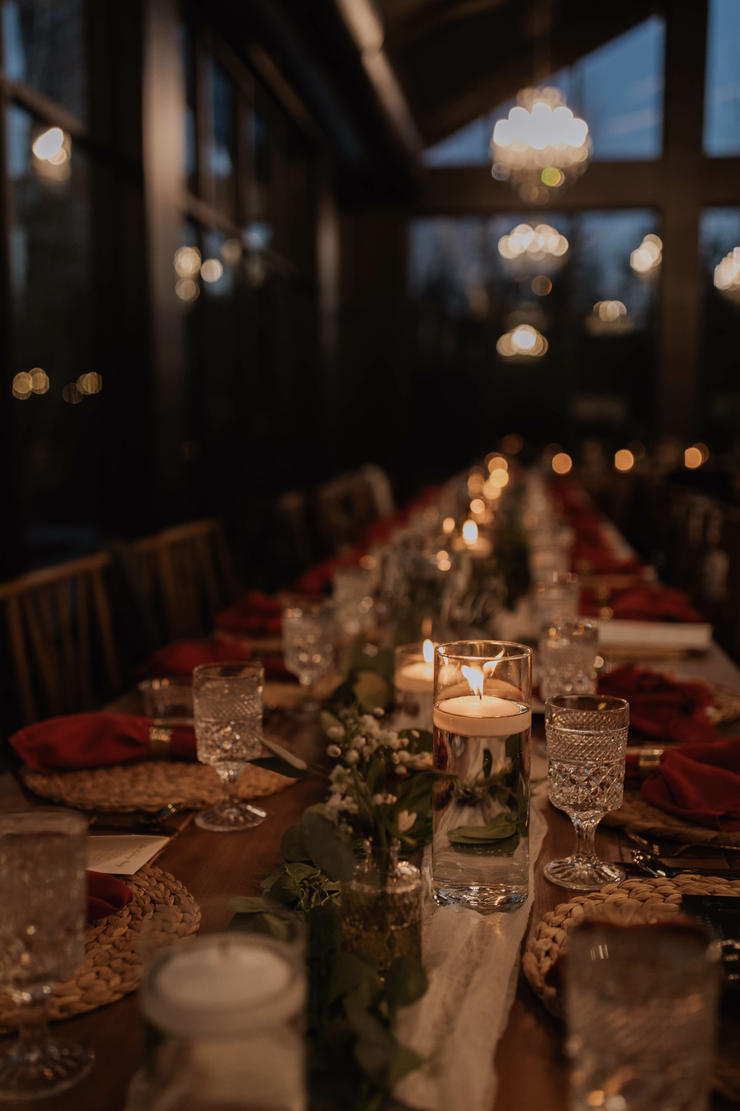 A close-up, warm-toned photograph of a long, beautifully styled reception table coordinated by Rooted Weddings & Events. The table runner features lush green eucalyptus and white floral garlands, accented by glowing floating candles in tall glass cylinders, ornate crystal glassware, woven chargers, and burnt orange napkins, set against a softly blurred background of evening windows and warm chandeliers.