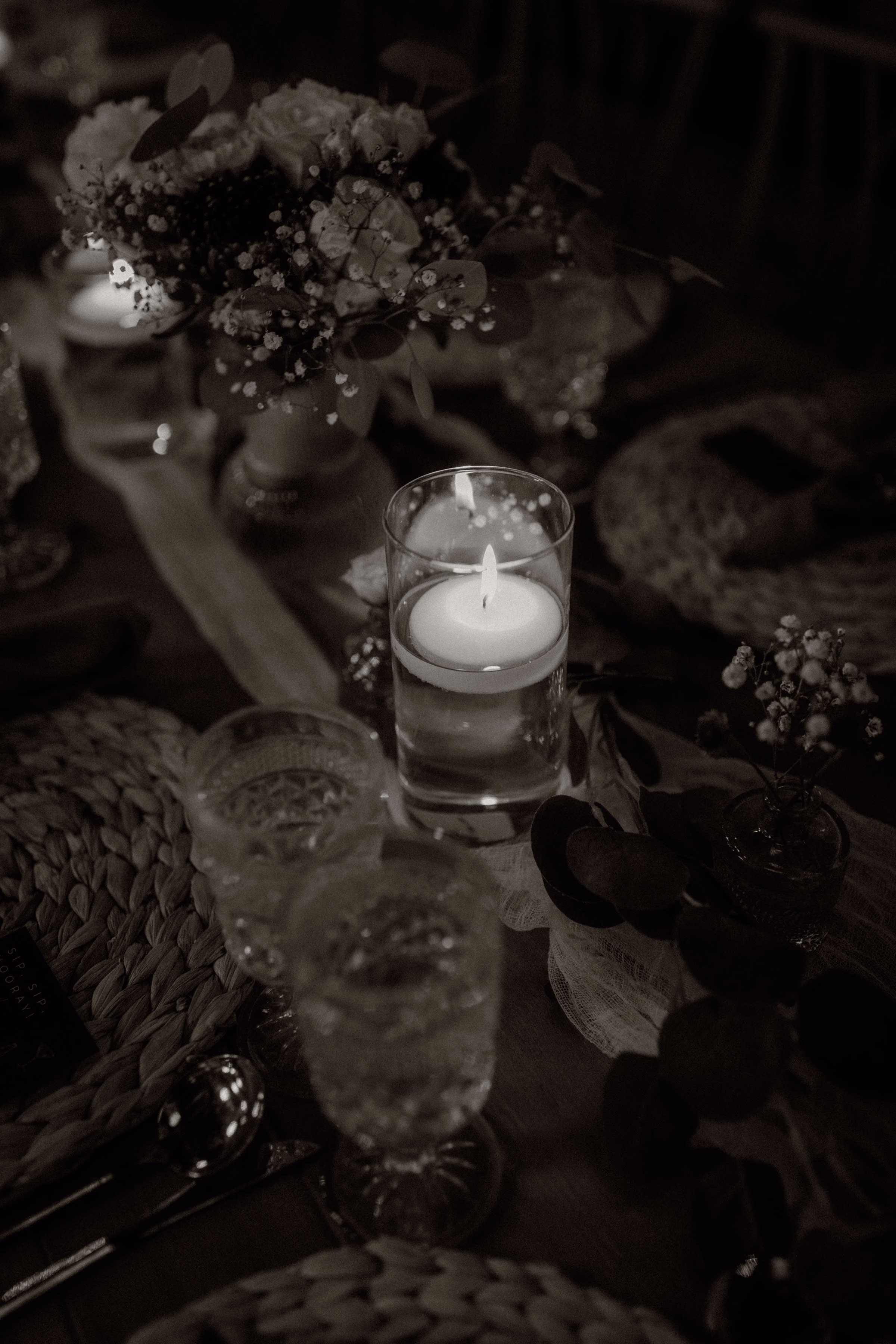 A black and white, moody overhead photograph highlighting an intimate reception table detail designed by Rooted Weddings & Events. A single lit floating candle glows inside a glass cylinder centerpiece, surrounded by delicate baby's breath, roses, and eucalyptus springs on a textured table runner, alongside woven placemats, formal silverware, and cut crystal glassware.