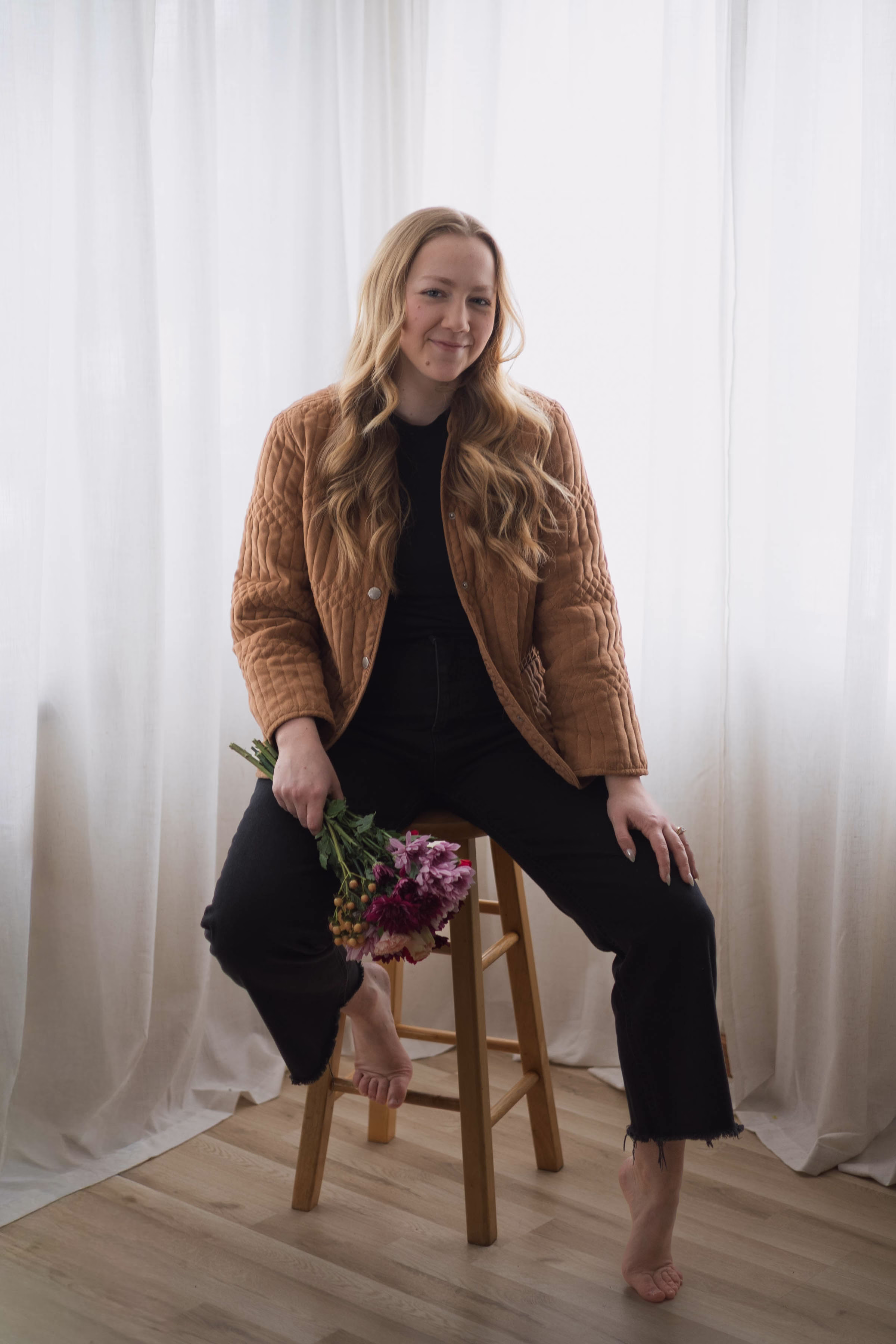 A professional portrait of Natalie Fisher, owner of Rooted Weddings & Events based out of La Crosse, Wisconsin. She sits barefoot on a wooden stool against a soft white curtain backdrop, smiling warmly at the camera. She wears a brown quilted jacket over a black top and black pants while holding a bouquet of dark pink flowers.