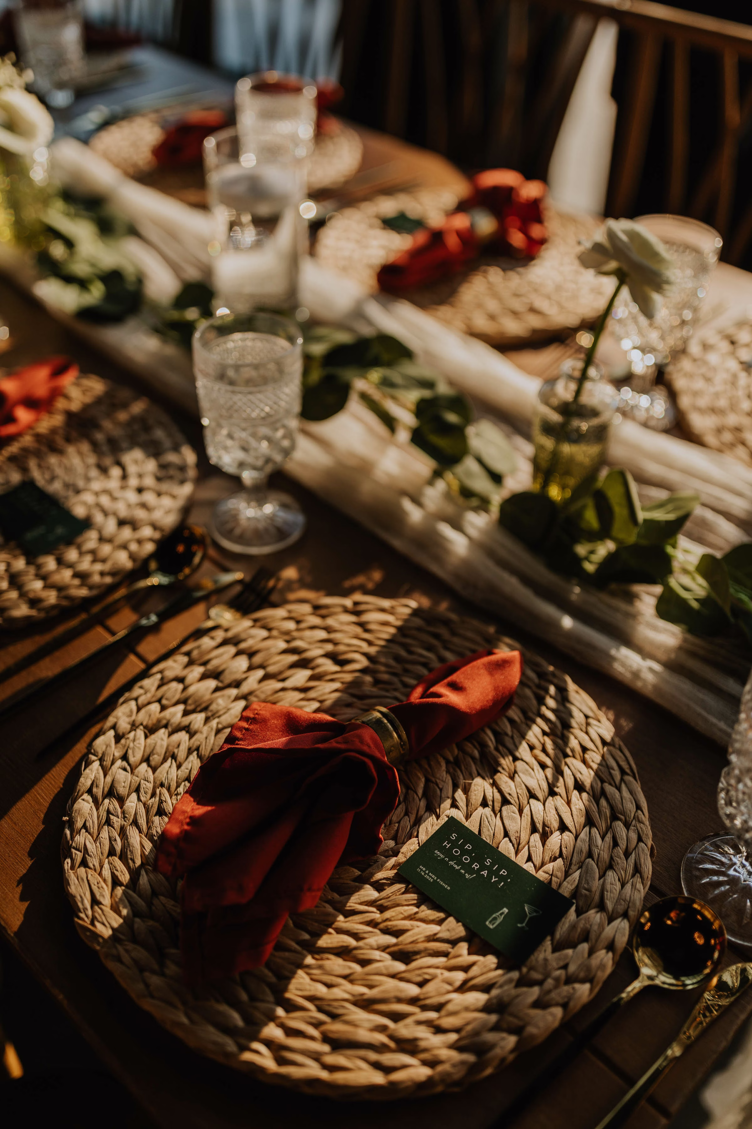 A warm, sunlit overhead photograph of a reception place setting styled by Rooted Weddings & Events. A round, woven straw charger mat holds a burnt orange napkin tied with a gold ring and a small dark green favor card that reads "SIP, SIP, HOORAY!", surrounded by cut crystal glassware, elegant gold flatware, and a flowing white gauze table runner adorned with fresh eucalyptus.