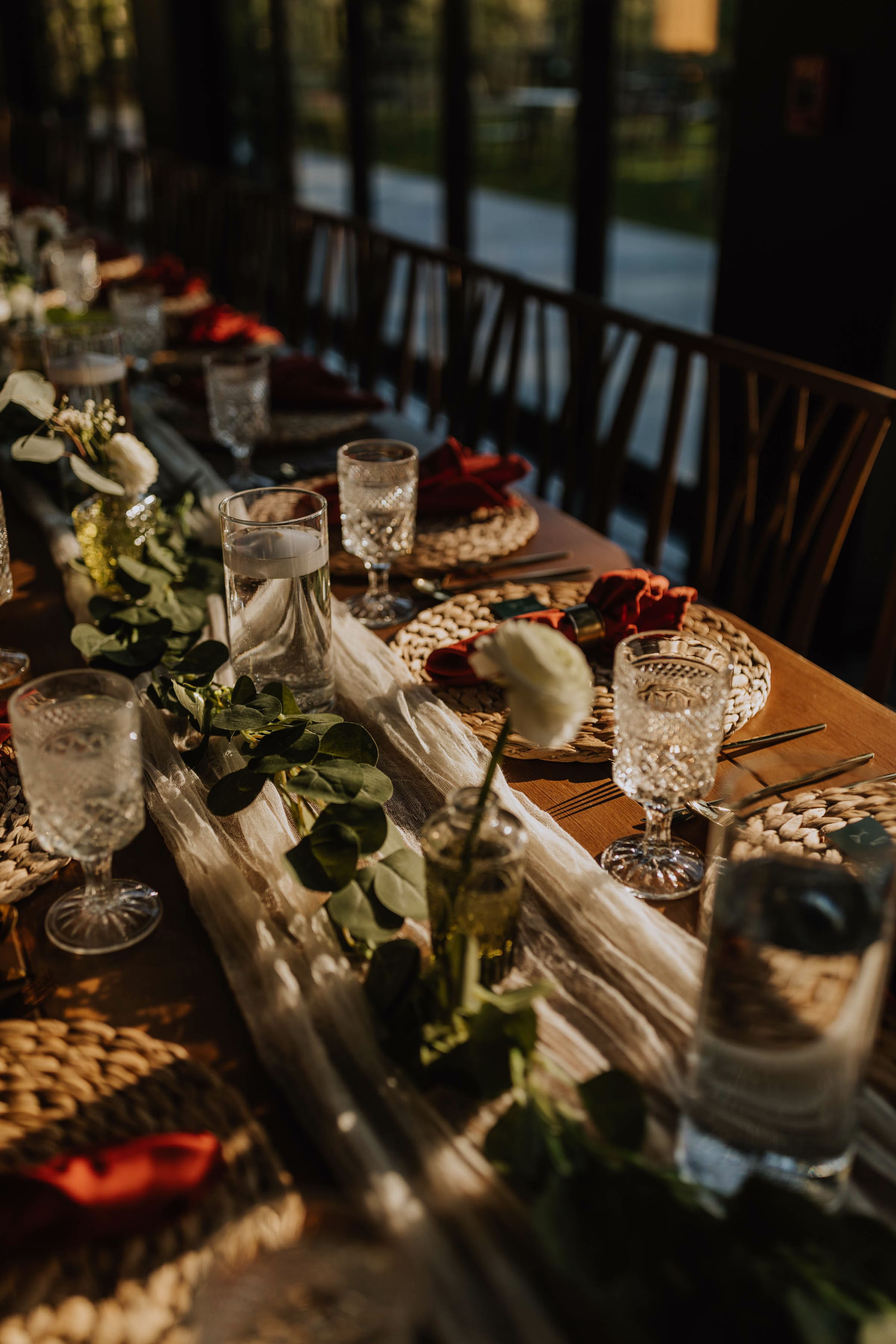 A warm, sun-drenched photograph looking down a long wedding reception table styled by Rooted Weddings & Events. A flowing white gauze runner stretches across the wooden table, woven with lush green eucalyptus branches, floating candles in tall glass cylinders, and a delicate single white flower in a small bud vase, alongside formal place settings featuring burnt orange napkins and elegant cut crystal glassware.