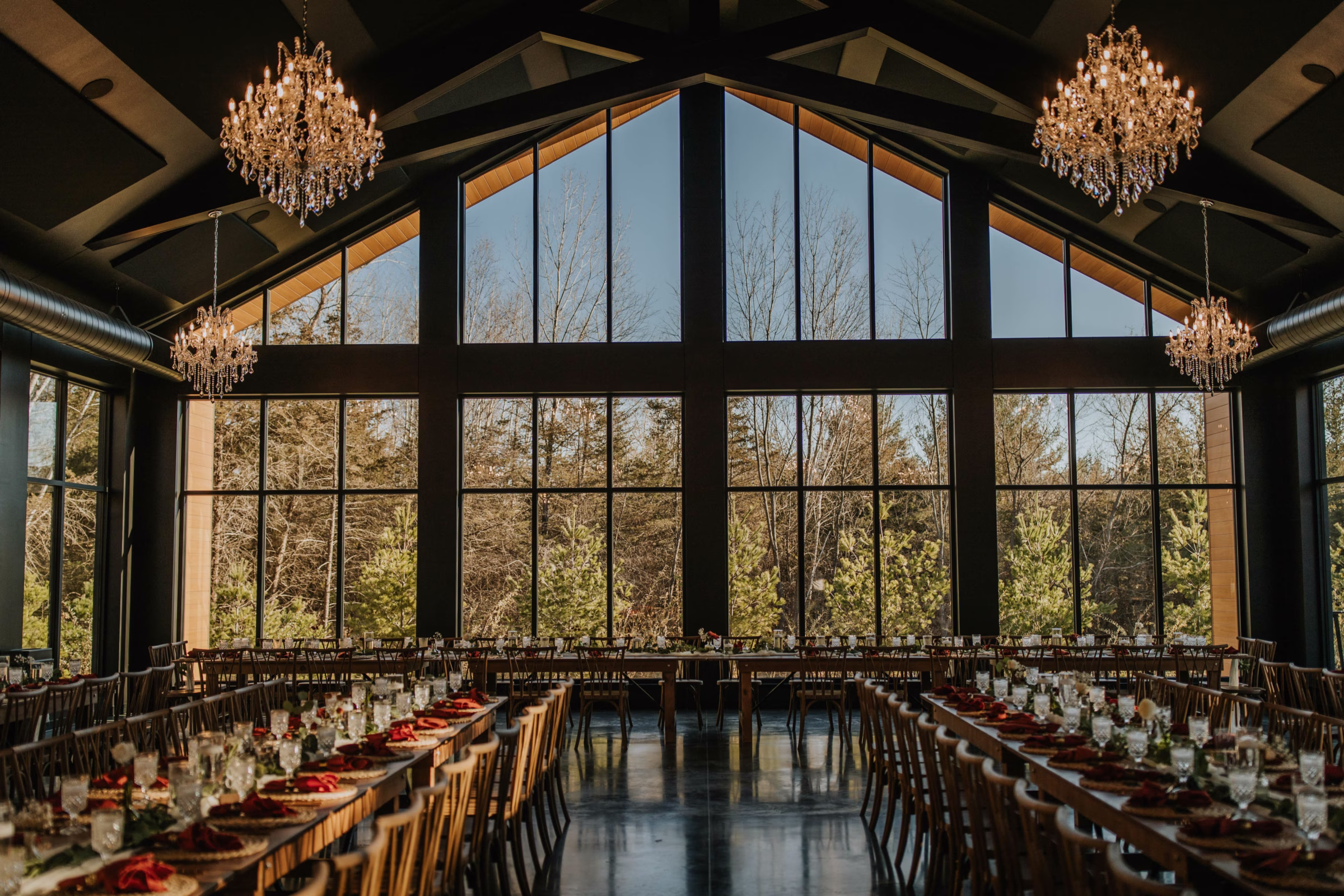 Large indoor wedding reception hall featuring a massive floor-to-ceiling A-frame window wall that looks out onto a dense evergreen forest under a clear blue sky. Elegant crystal chandeliers hang from dark wood vaulted ceilings, illuminating long reception tables set with burnt orange napkins, glassware, and floral arrangements. Designed by Rooted Weddings & Events