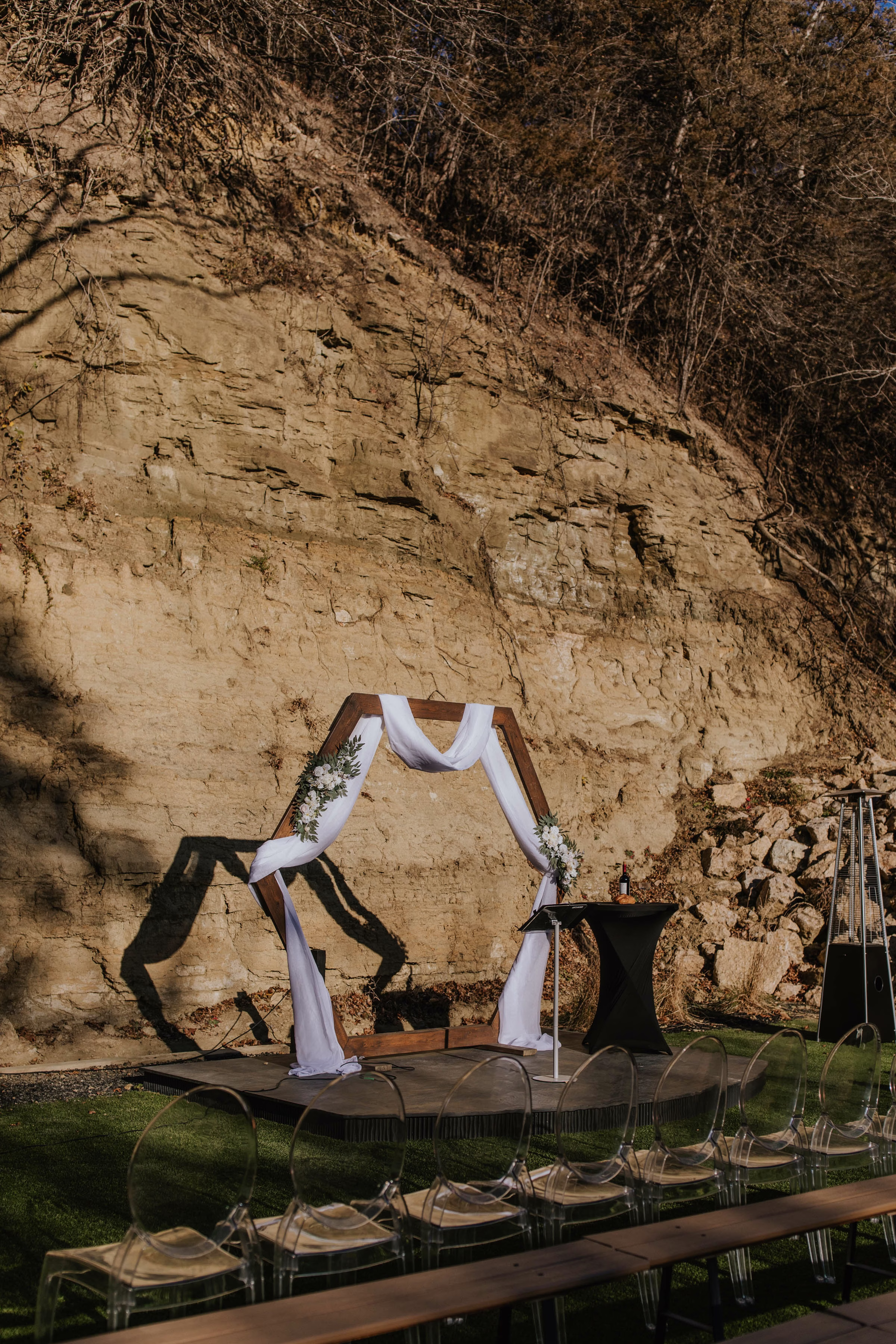 A warm, outdoor wedding ceremony setup styled by Rooted Weddings & Events. A wooden hexagonal arch draped in white fabric and adorned with white floral arrangements stands on a dark stage against a dramatic, high sandstone bluff backdrop, with rows of modern clear ghost chairs arranged on the lawn in front.