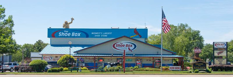 The exterior storefront of The Shoe Box in Black Earth, WI, famously known as the Midwest’s largest shoe store. The large retail building features prominent red, white, and blue signage, a statue of a monkey on the roof, an American flag, and a "Welcome to Black Earth" sign.