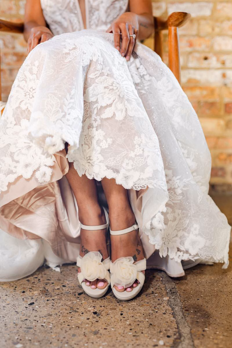 A bride in an intricate lace gown shows off her elegant ivory bridal heels featuring delicate mesh flower detailing on the toe straps, purchased from the massive footwear selection at The Shoe Box in Black Earth, WI.