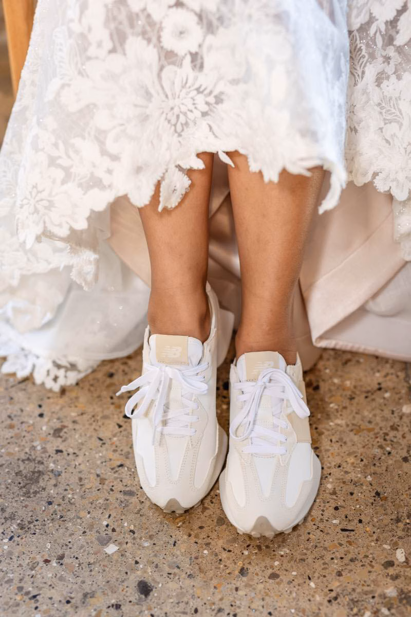 A bride in a lace gown lifts her dress to reveal classic cream and white New Balance 327 lifestyle sneakers, showcasing stylish and comfortable bridal sneaker options available at The Shoe Box in Black Earth, WI.