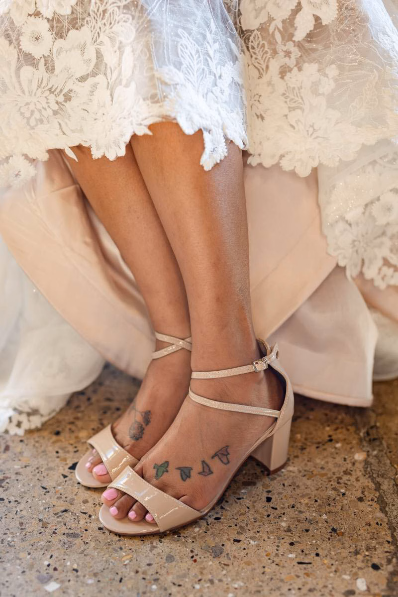 A bride in a lace gown displays her nude patent leather block heel sandals with crisscross ankle straps, showcasing elegant special occasion and bridal footwear available at The Shoe Box in Black Earth, WI.