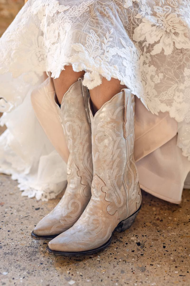A bride in a lace wedding gown shows off her distressed cream and beige Dan Post cowgirl boots with intricate western embroidery, showcasing bridal and western footwear options from The Shoe Box in Black Earth, WI.